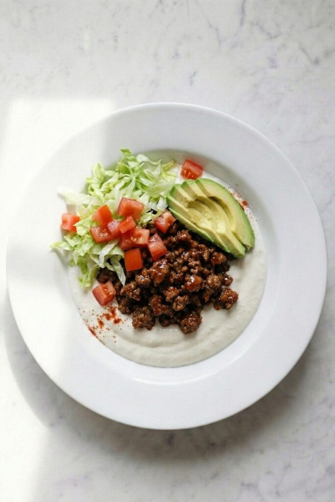 Close-up of an assembled taco dip bowl showing triangulation of toppings: shredded lettuce, diced tomatoes, and sliced avocado over seasoned beef and whipped base.