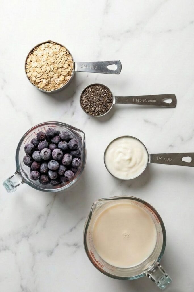 Overhead flat lay on white marble showing 3/4 cup rolled oats, 1 tbsp chia seeds, frozen blueberries, and oat milk for meal prep.