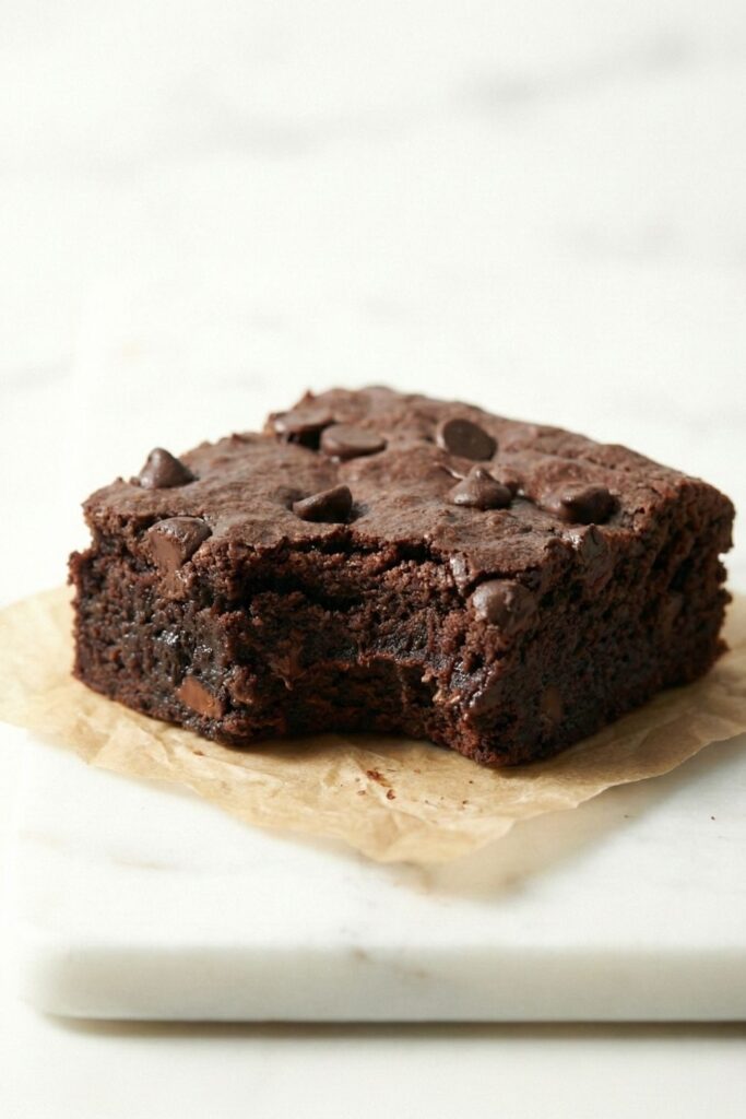 A single fudgy brownie on crumpled parchment paper with a bite taken out, showing the dense crumb structure.