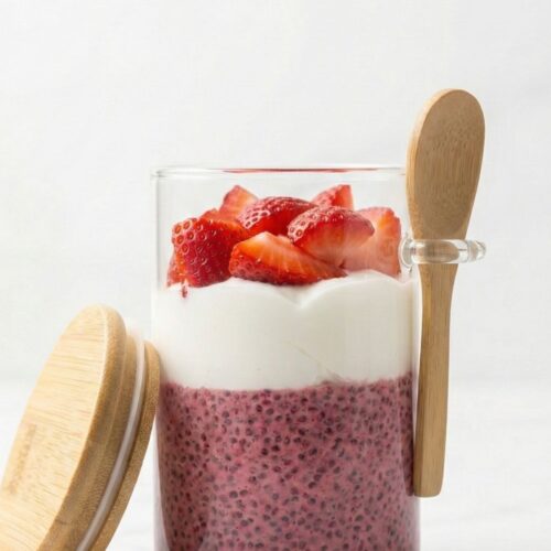 High protein raspberry chia pudding parfait in a glass jar with spoon holder, layered with greek yogurt and fresh berries on a white marble background.
