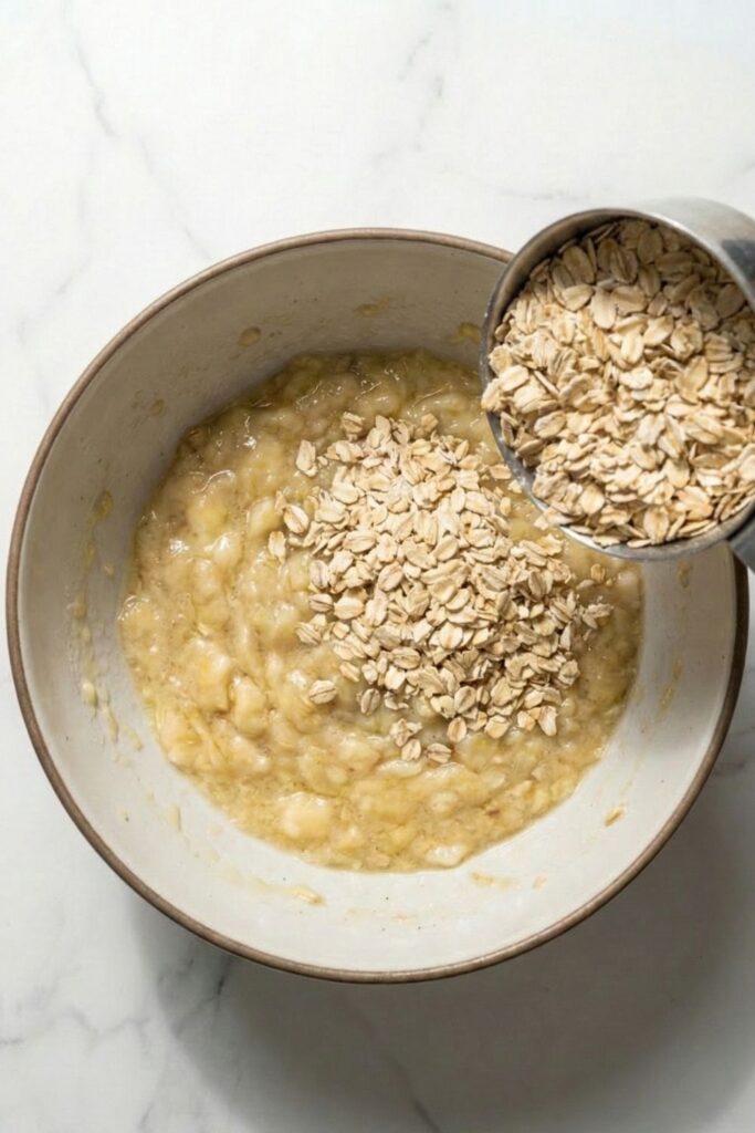 Action shot pouring exactly 1 ½ cups of dry old-fashioned rolled oats into 1 ½ cups of wet mashed banana to create a high-fiber cookie dough base without the use of eggs or flour.