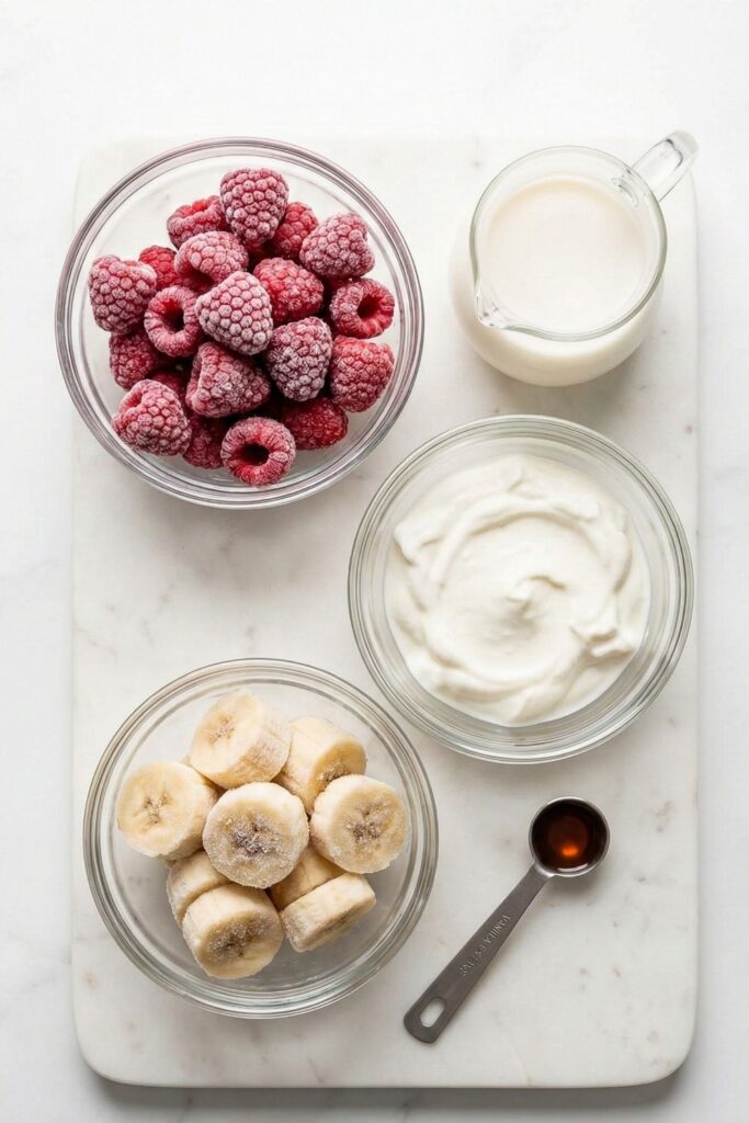 Top-down flat lay of frozen raspberries, sliced bananas, and thick Greek yogurt in clear glass bowls on a white marble board, with a small pitcher of milk and vanilla extract.