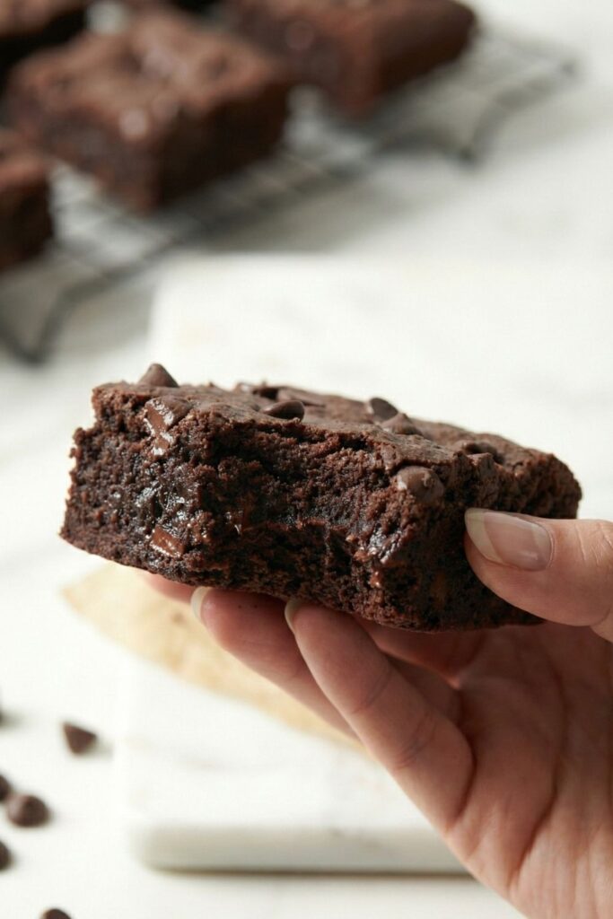 A hand holding a rich chocolate brownie close to the camera to demonstrate the serving size and moist texture.