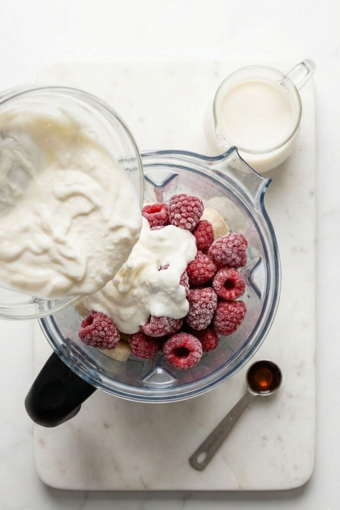 Overhead view of creamy Greek yogurt being poured into a blender canister filled with frozen red raspberries and banana slices on a white marble surface.