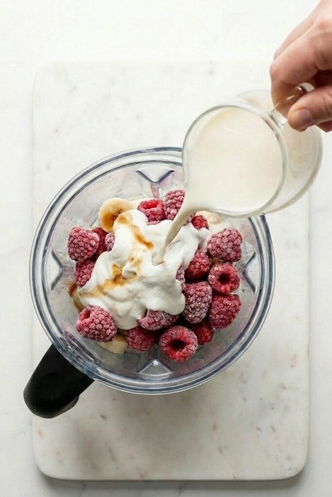Action shot of almond milk being poured from a clear glass pitcher into a blender, cascading over greek yogurt and frozen berries to show liquid consistency.
