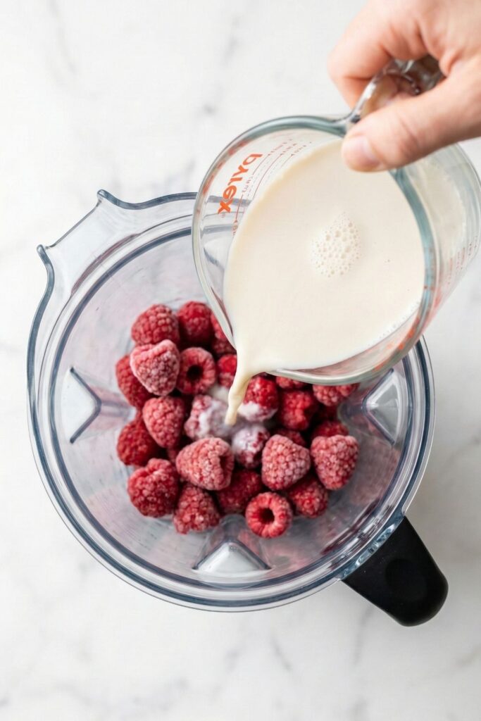 Pouring oat milk into a blender containing frozen raspberries to create the pink milk base for the chia pudding.