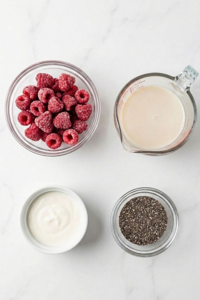 Overhead flat lay of ingredients for raspberry chia pudding, including frozen raspberries, oat milk, vanilla yogurt, and chia seeds arranged on a white marble counter.