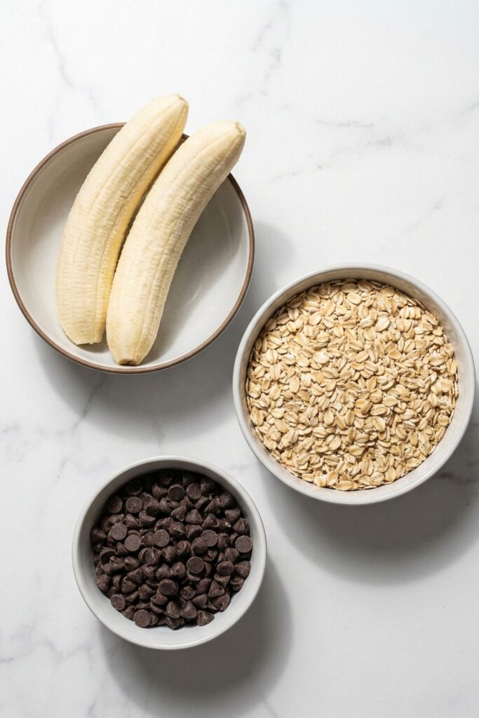 Overhead shot of raw ingredients for 3-ingredient banana oatmeal cookies on a white marble surface, featuring 2 large ripe bananas, exactly 1 ½ cups of old-fashioned rolled oats, and ⅓ cup of chocolate chips.