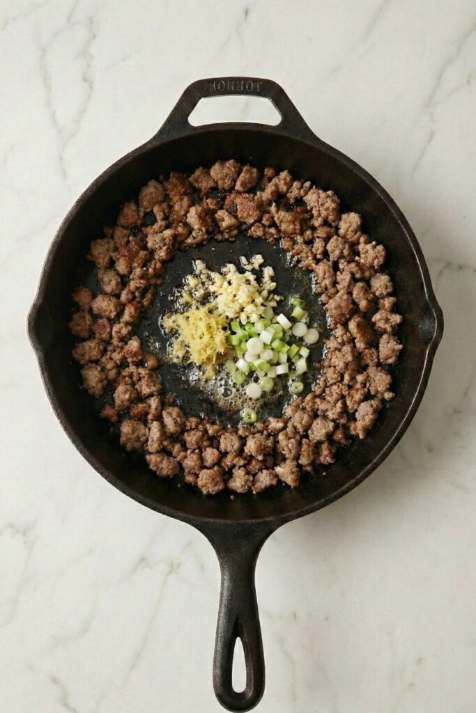 Minced garlic, grated ginger, and scallion whites sizzling in hot oil in the center of a skillet surrounded by cooked ground turkey.