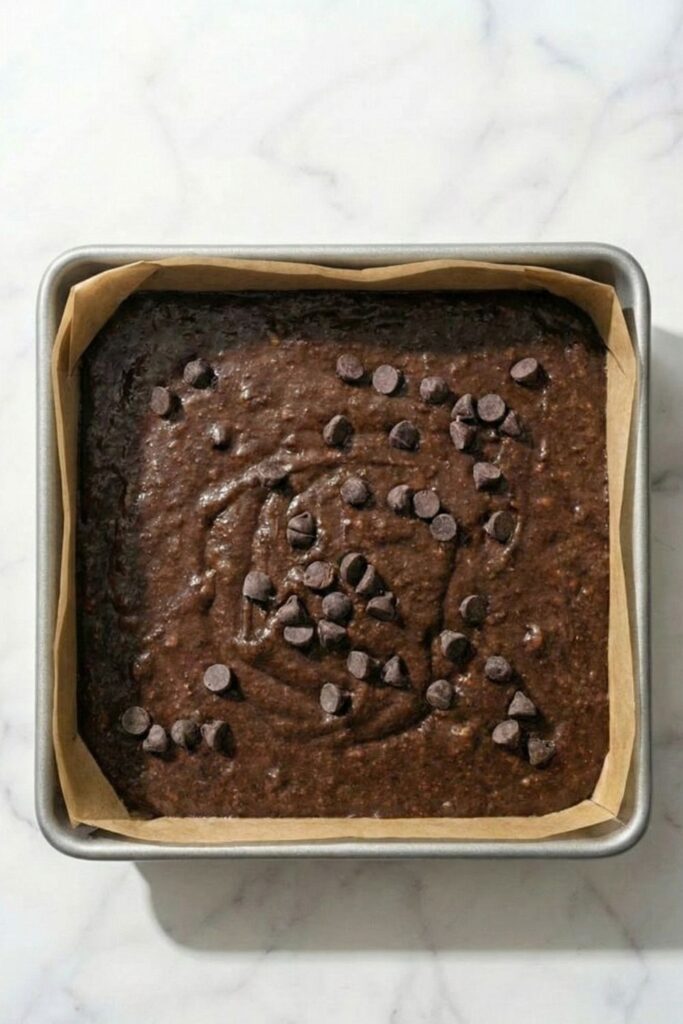 Smooth, raw 3 ingredient brownie batter spread completely flat into an 8x8 inch square baking pan lined with overhanging parchment paper.