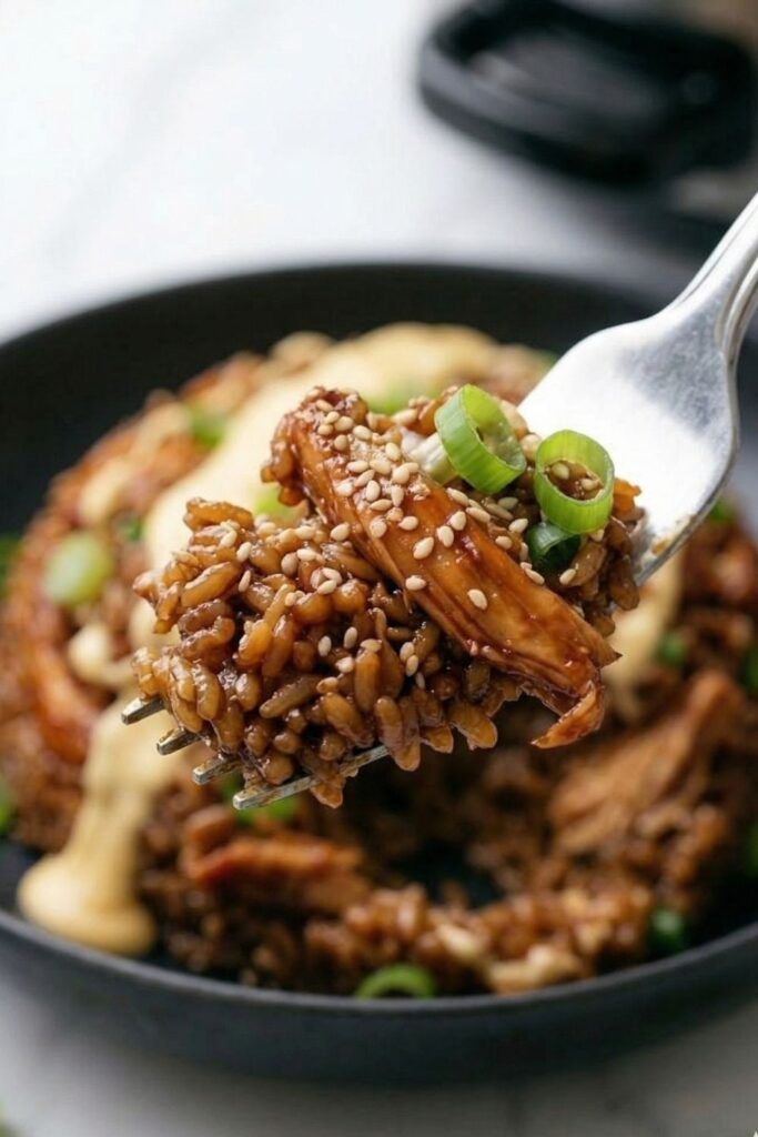 Close-up eye-level view of a fork lifting a glistening bite of shredded teriyaki chicken and sticky short-grain rice, showcasing the thick glaze and distinct rice grains against a blurred background.